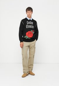 Jonge man in een zwart sweater met de tekst "London Chronicles" en een rode roos, beige cargobroek, blauwe shirt en tan sneakers, staand.