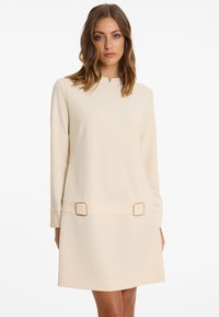 Beige Langarmkleid mit geradem Schnitt, feinem Ausschnitt, zwei quadratischen Schnallen in der Taille und glattem, strukturiertem Stoff.