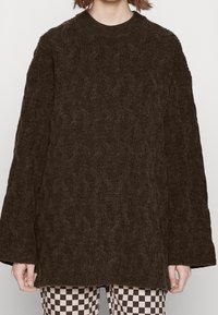 Brauner Strickpullover mit hohem Kragen und weiten Ärmeln, der ein strukturiertes Muster aufweist. Kombiniert mit karierter Hose.