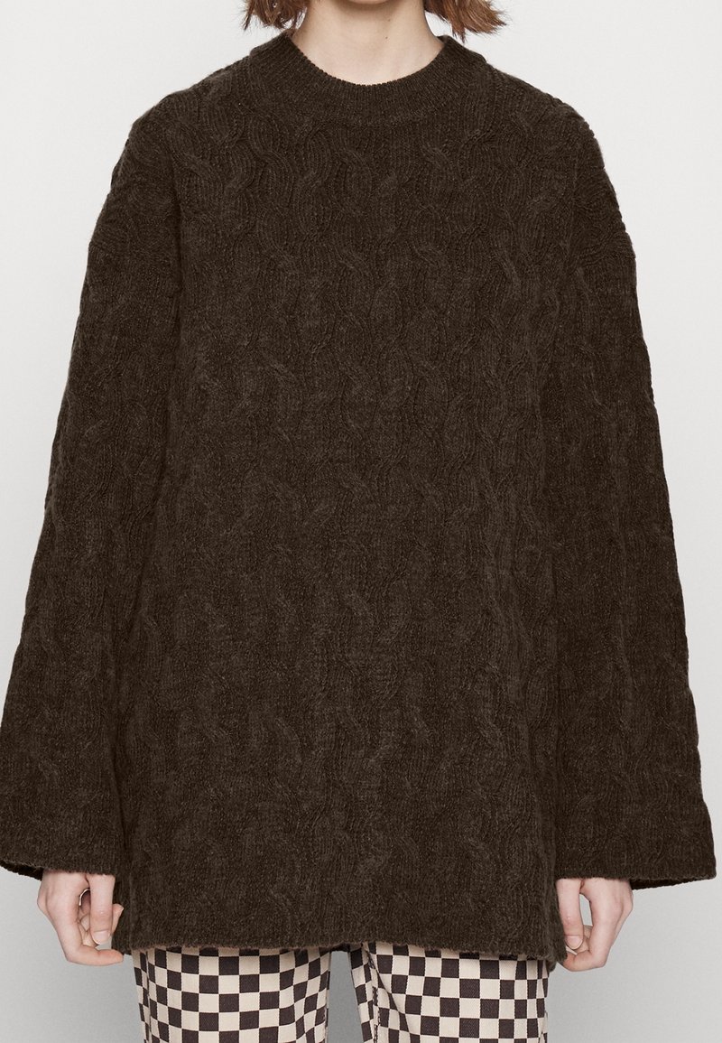 Brauner Strickpullover mit hohem Kragen und weiten Ärmeln, der ein strukturiertes Muster aufweist. Kombiniert mit karierter Hose.