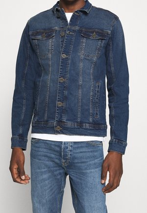 Homme portant une veste en denim bleu ajustée boutonnée sur une chemise blanche, associée à un jean denim bleu clair sur un fond uni.