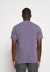Homme debout de dos, portant un t-shirt violet à manches courtes et un pantalon gris foncé sur un fond clair uni.