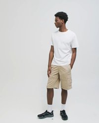 Tânăr care poartă un tricou alb, pantaloni scurți cargo bej, șosete albe și adidași negri, stă în picioare și privește într-o parte, pe un fundal simplu.