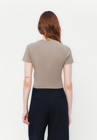 Žena s dlouhými červenými vlasy, oblečená v krátkém taupe crop topu s krátkým rukávem a černých kalhotách s vysokým pasem, otočená zády k bílému pozadí.