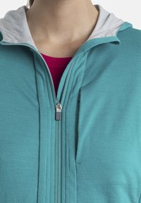 Felpa con zip color teal, foderata in grigio e dotata di due zip frontali. Tessuto morbido con design aderente, indossata sopra una maglietta rosa.