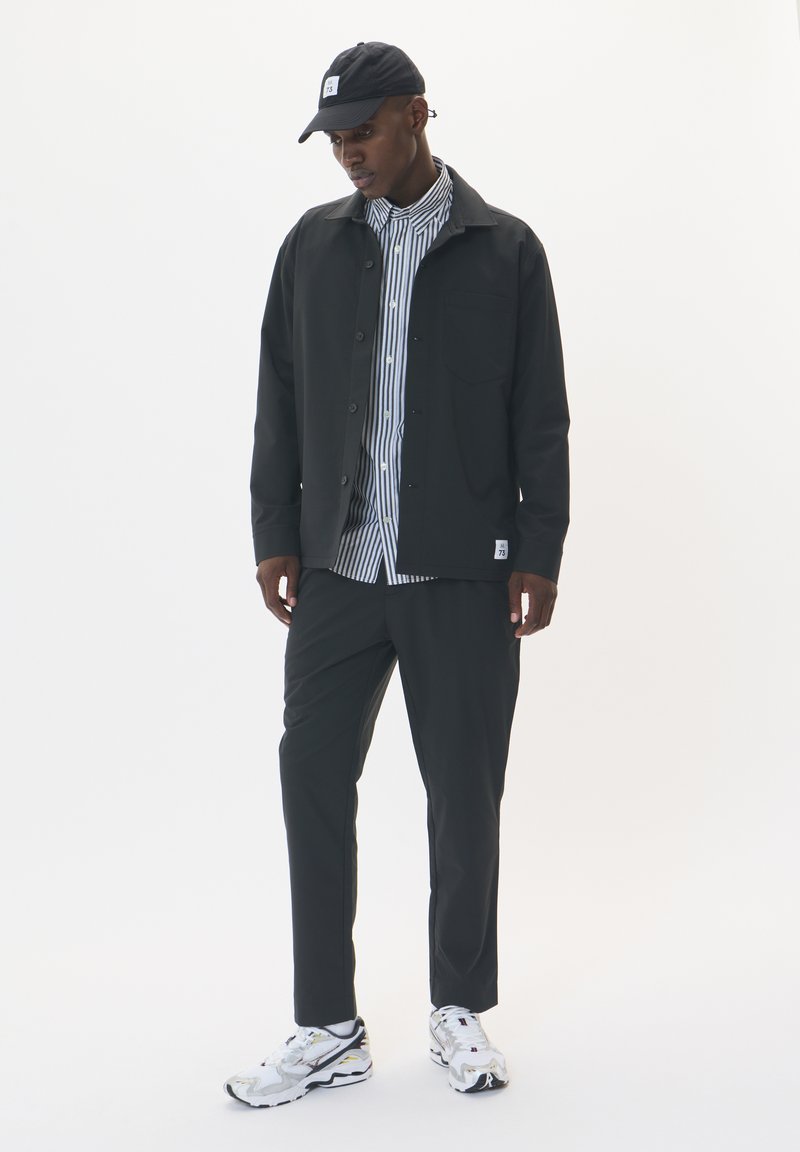 Czarna kurtka zapinana na guziki nałożona na pasiastą koszulę, w połączeniu z czarnymi spodniami i białymi trampkami z żółtymi akcentami. Czapka dopełnia całość.