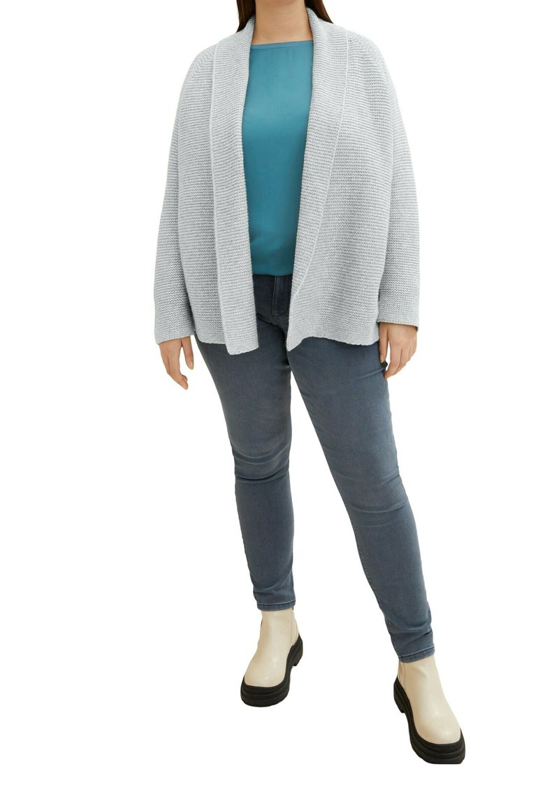 Cardigan grigio chiaro con frontale aperto e design testurizzato, indossato su una maglietta teal, abbinato a jeans grigio scuro e stivaletti beige con tacco platform.