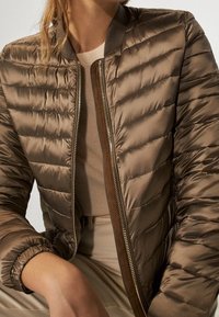 Gevoerde bomberjack in donker taupe met een ritsluiting, ribbelkraag en -manchetten, voorzien van een chevronpatroon en een gladde, glanzende textuur.