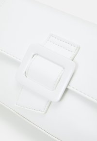 Biała kopertówka z ekoskóry z kwadratową klamrą. Charakteryzuje się gładką, elegancką fakturą oraz subtelnym przeszyciem. Kompaktowy, prostokątny kształt.