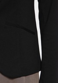 Blazer negro hecho de tela texturizada con corte ajustado, que presenta un bolsillo lateral y un dobladillo redondeado.