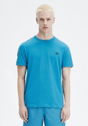 Jonge man met kort blond haar, gekleed in een felblauw T-shirt met korte mouwen en bijpassende korte broek, staand tegen een effen witte achtergrond.