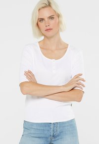 Weißes, geripptes Kurzarmshirt mit einem Rundhalsausschnitt und einer Knopfleiste mit vier Knöpfen. Der Stoff ist glatt und körperbetont. Kombiniert mit hellblauen Jeans.