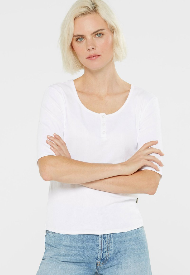 Weißes, geripptes Kurzarmshirt mit einem Rundhalsausschnitt und einer Knopfleiste mit vier Knöpfen. Der Stoff ist glatt und körperbetont. Kombiniert mit hellblauen Jeans.