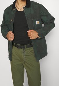 Veste en coton vert avec un col en velours côtelé noir, poches avant et patch de marque ; portée sur un haut noir et associée à un pantalon vert olive.