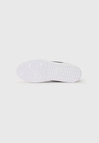 A sola do sapato apresenta um design em borracha branca com padrões de textura para aderência e acentos circulares; o lado inclui um emblema circular e detalhes da marca.