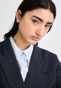 Blazer blu navy con rever affilati su una camicia azzurro chiaro, caratterizzato da una texture liscia e dettagli con bottoni, indossato da un modello con capelli scuri.