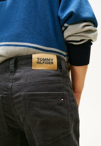 Persoon in donkere corduroy Tommy Hilfiger broek met zichtbaar logopatch en een blauw-beige gestreepte trui met opgerolde mouwen.