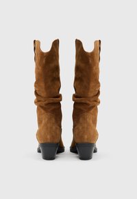 Botas de ante marrón con un diseño holgado, puntas afiladas y un tacón apilado. Presentan costuras laterales curvas y una textura suave.