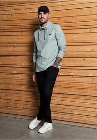 Světle šedá mikina s kapucí a šňůrkami, černé kalhoty a bílé tenisky. Oblečení má malé logo na hrudi a hladkou texturu.