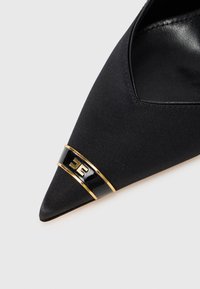 Black canvas pointed-toe shoe with a gold and black shiny accent band featuring a logo. Smooth texture on the inner side.