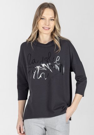 Kobieta o jasnych, falowanych włosach ubrana w czarną, długorękawową bluzkę z srebrnym napisem "la dolce vita" i jasnoszare spodnie, stoi z jedną ręką w kieszeni.