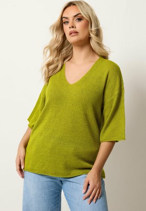 Blonde Frau mit losen Wellen, die einen limettengrünen, strukturierten V-Ausschnittpullover mit ellbogenlangen Ärmeln und hellblaue Jeans trägt.