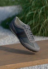 Zapatilla deportiva gris que presenta una combinación de materiales lisos y texturizados, con un diseño de cordones y acentos a rayas en contraste.
