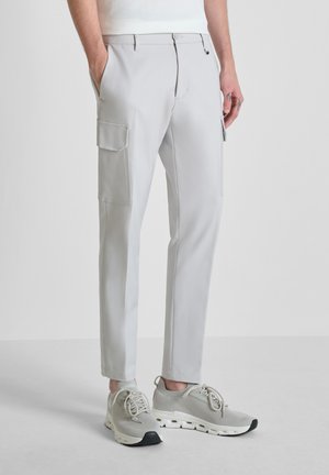 Hombre con pantalones cargo gris claro de corte ajustado con bolsillos con solapa y zapatillas deportivas gris claro, de pie con las manos parcialmente en los bolsillos.