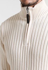 Beige ribbade zip-up tröja med hög krage, utrustad med en silverfärgad dragkedja och svart dragbane, tillverkad av mjukt, texturerat tyg.