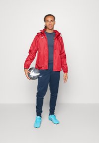 Veste légère rouge avec une capuche, assortie à une chemise à manches longues bleu marine et un pantalon de survêtement. Chaussures de football bleues, tenant un ballon noir et blanc.