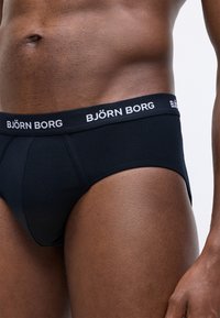 Detailný záber na osobu v čiernych trenkách Björn Borg, zobrazený pás, bok a horné stehno na jednofarebnom pozadí.