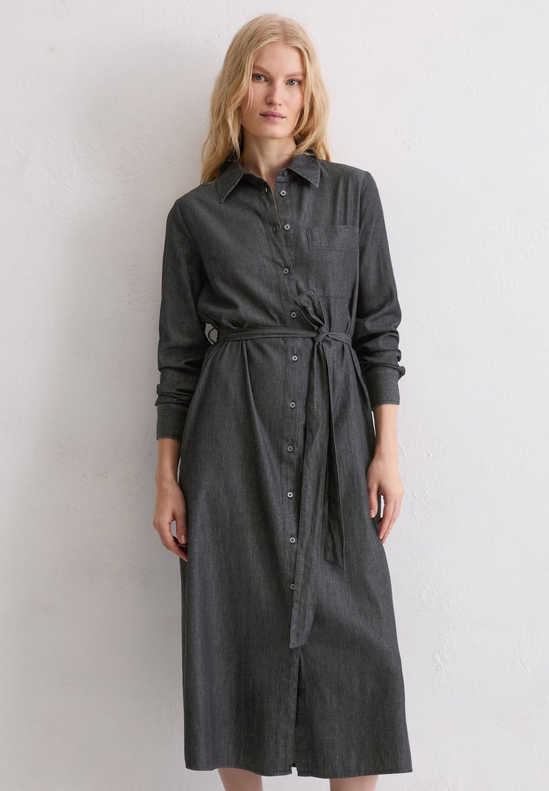 Szara sukienka koszulowa z denimu z paskiem do wiązania, długimi rękawami, zapięciem na guziki i kieszenią na piersi. Tkanina ma miękkie, lekko teksturowane wykończenie.