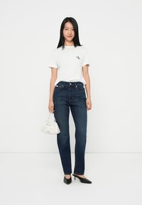 T-shirt blanc à manches courtes avec logo, jeans taille haute bleu marine, escarpins noirs pointus et petit sac texturé blanc. Fond uni.