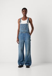 Jeansoverall med ljusblå tvätt, vida ben, justerbara axelband, framficka och silverfärgad metall. Bärs över en vit tanktop.