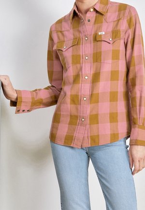 Personne portant une chemise à carreaux rose et marron avec poches poitrine, associée à un jean bleu clair, debout devant un fond uni.