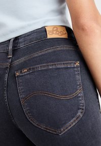 Jeans de mezclilla oscura con un diseño ajustado, que presentan una etiqueta de cuero marrón y costuras naranjas en contraste en los bolsillos y las costuras.