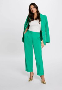 Blazer verde su misura e pantaloni coordinati, realizzati in tessuto liscia. I pantaloni hanno delle pince e risvolti. Un top bianco e dècolletè dorate completano il look.