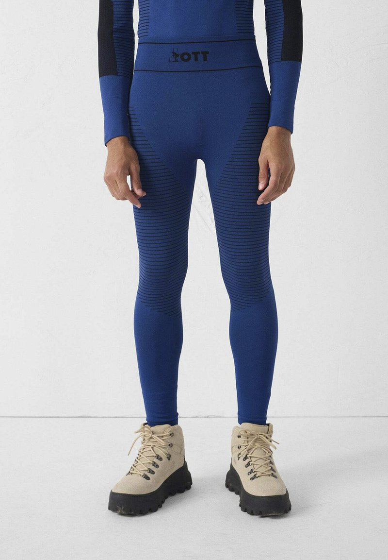 Blauwe sportleggings met getextureerde strepen en een zwarte tailleband met het logo "ZOTT." Gecombineerd met beige wandelschoenen.