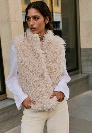 Ung kvinde med krøllet brunt hår iført en lys beige fluffy vest over en hvid langærmet skjorte og cremefarvede bukser, står udendørs.