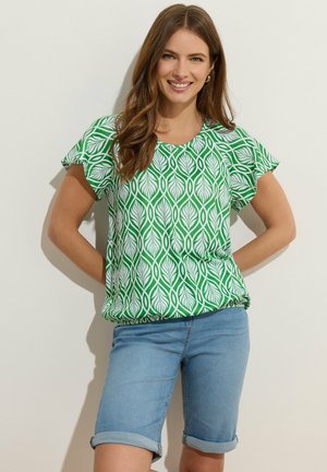 Nainen hymyilee, hänellä on pitkät ruskeat hiukset ja päällään vihreä-valkoinen lehtikuosinen lyhythihainen paita sekä käärityt siniset farkkushortsit.