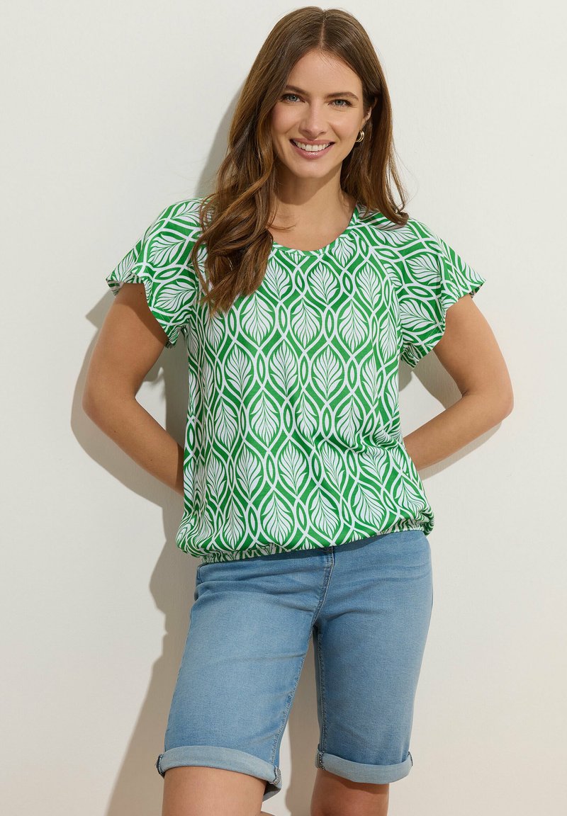 Vrouw glimlachend met lang bruin haar, gekleed in een groen-wit topje met bladerenpatroon en opgerolde blauwe denim korte broek.