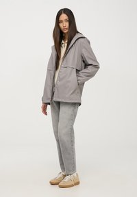 Szara wodoodporna kurtka z kapturem, dwoma przednimi kieszeniami, luźny krój, w połączeniu z jasnoszarymi dżinsami i beżowymi sneakersami.