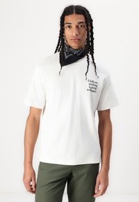Weißes Baumwoll-T-Shirt mit schwarzem Drucktext auf der Brust. Das Model trägt ein schwarzes Bandana und olivgrüne Hosen. Lässige Passform.