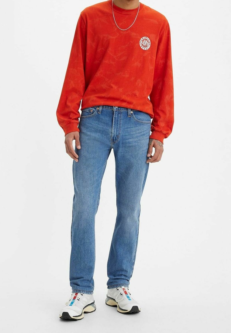 Röd långärmad skjorta med en grafisk logotyp, kombinerad med blå denimjeans. Skorna är vita sneakers med färgglada detaljer och en strukturerad sula.