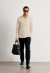 Beige langermet polo-skjorte med glidelås, svarte bukser, svart lærveske, solbriller, går på jevn grått gulv mot en ensfarget bakgrunn.