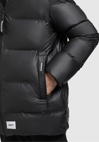 Schwarze Steppjacke mit matter Oberfläche, ausgestattet mit einer Reißverschlusstasche, horizontal gestepptem Design und Logo-Tag am Saum.