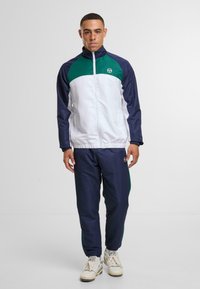 Tuta sportivo in blu, verde e bianco. La giacca presenta un collo alto, chiusura con zip e polsini elastici. I pantaloni hanno strisce laterali e una vita elastica.
