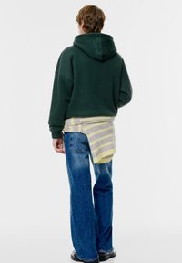 Felpa verde con grande grafica sul retro, indossata sopra un maglione leggero a righe. Abbinata a jeans blu larghi e calzature scure. Design semplice.