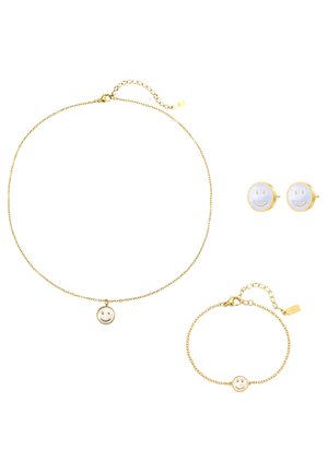 Frauenhandgelenk mit einem goldenen Kettenarmband mit rundem Smiley-Anhänger, trägt ein weißes, langärmeliges, geripptes Oberteil.