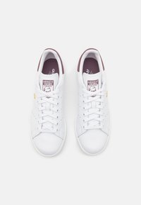 Zapatillas de cuero blancas con un diseño de corte bajo, acentos burdeos en la lengüeta y el talón, que presentan una suela de goma texturizada y seis ojetes.